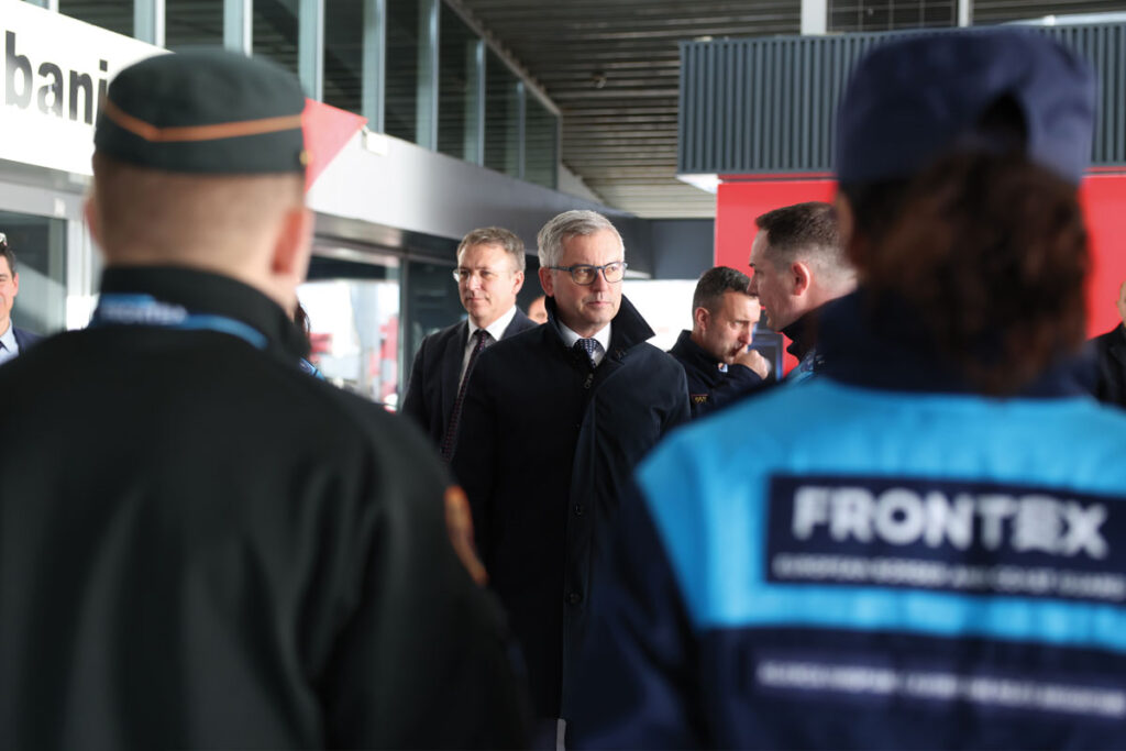 Im Vordergrund sind unscharf zwei Polizist*innen zu sehen, eine von ihnen trägt eine Frontext-Uniform. Im Hintergrund ist der EU-Kommissar Magnus Brunner zu sehen. Es handelt sich um ein Foto einer Visite der Grenzkontrolle im Hafen von Durrës in Albanien.